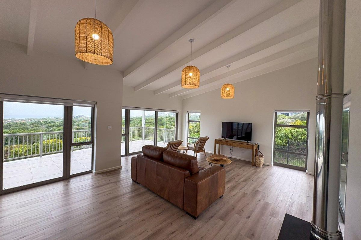 Spacious living room featuring vaulted ceilings with exposed beams, wood-look flooring, a modern freestanding fireplace, and large sliding doors opening onto a scenic balcony.
