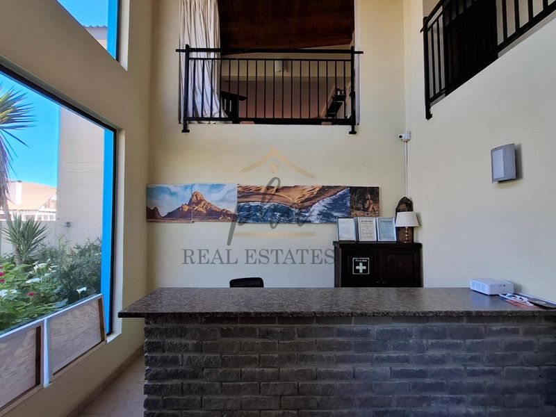 Double-volume reception area featuring a brick-fronted desk, large windows providing natural light, and an overlooking mezzanine level with metal railings.