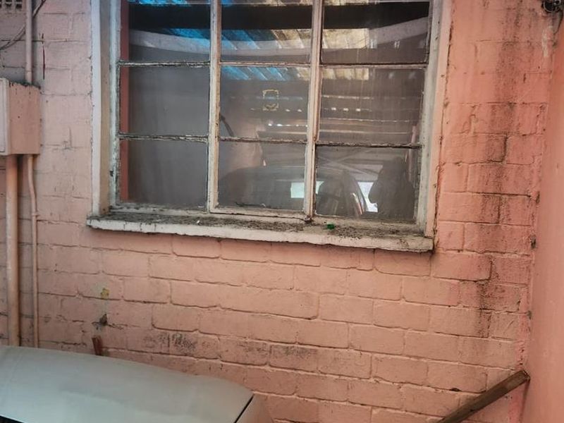 An exterior rear area featuring a weathered brick wall, a steel-framed window, and corrugated roof sheeting showing signs of significant wear and deterioration.
