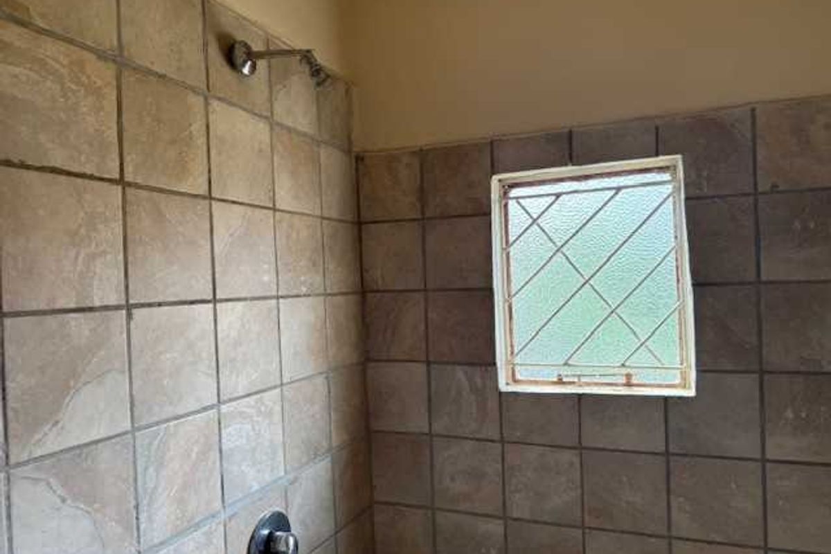 A dated shower enclosure featuring brown square wall tiles, a fixed shower head, and a small window with security bars.