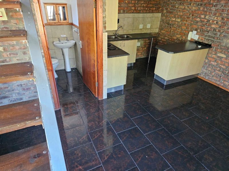 Open plan living area featuring exposed brick walls, dark tiled flooring, a compact kitchen with built-in cabinetry, and an adjacent bathroom with a pedestal basin.