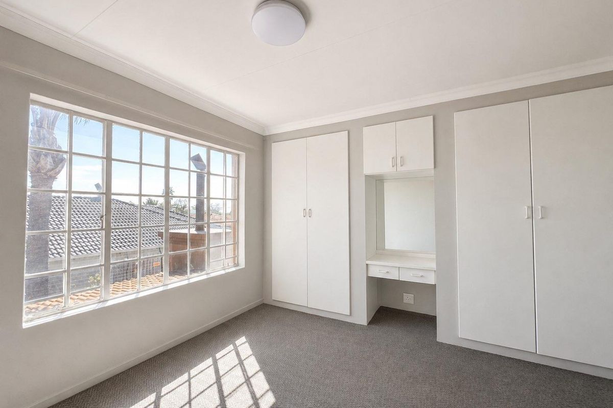 A bright, empty bedroom featuring neutral carpeting, large grid-style windows, and extensive built-in cabinetry with an integrated vanity area.
