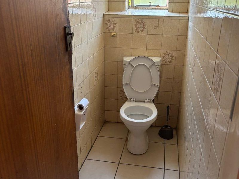 A compact, dated bathroom featuring ceramic wall and floor tiles, a standard toilet, and a window for natural light.