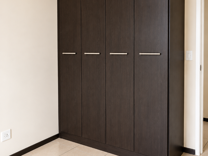 A clean, empty bedroom featuring a modern dark wood built-in wardrobe and neutral tiled flooring.