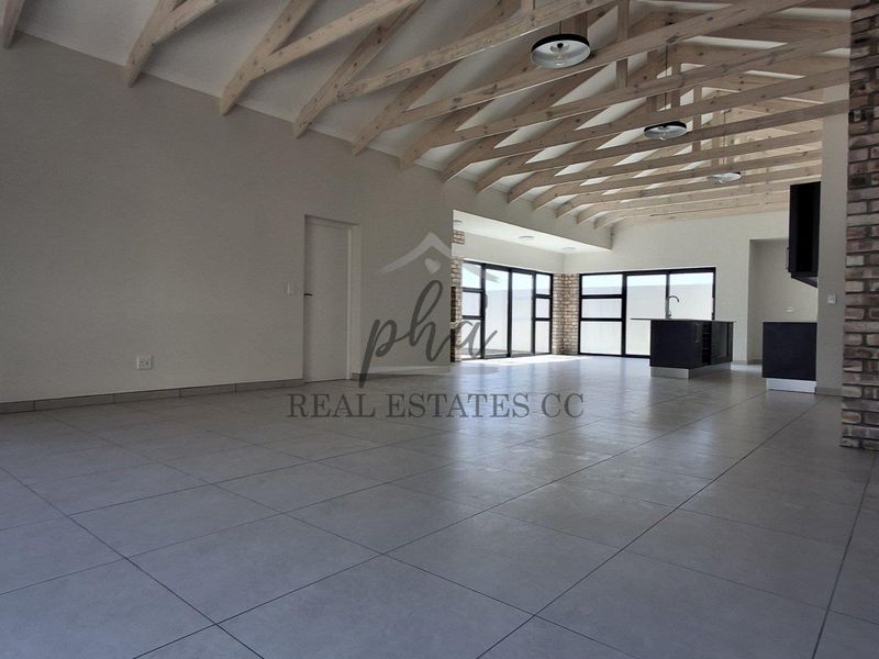 Spacious open-plan living area featuring exposed timber roof trusses, a rustic brick feature wall, modern tiled flooring, and a sleek kitchen island.