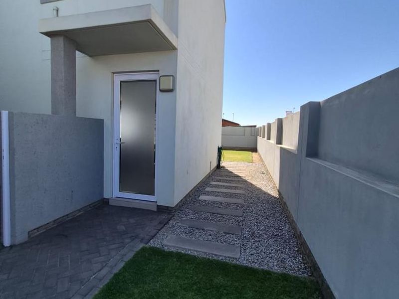 A side view of the townhouse exterior featuring a paved walkway, gravel path, boundary wall, and a frosted glass entrance door.