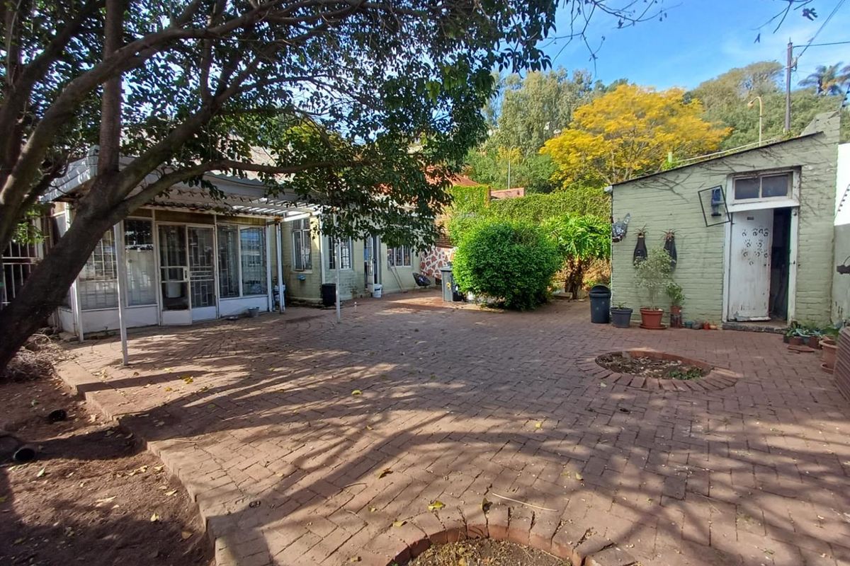 A spacious rear courtyard featuring brick paving, a covered patio area, and a separate brick outbuilding set within a mature garden.