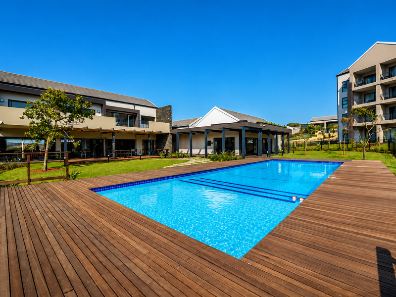 A well-maintained outdoor pool area featuring extensive timber decking, landscaped gardens, and modern apartment buildings with covered patios and balconies.