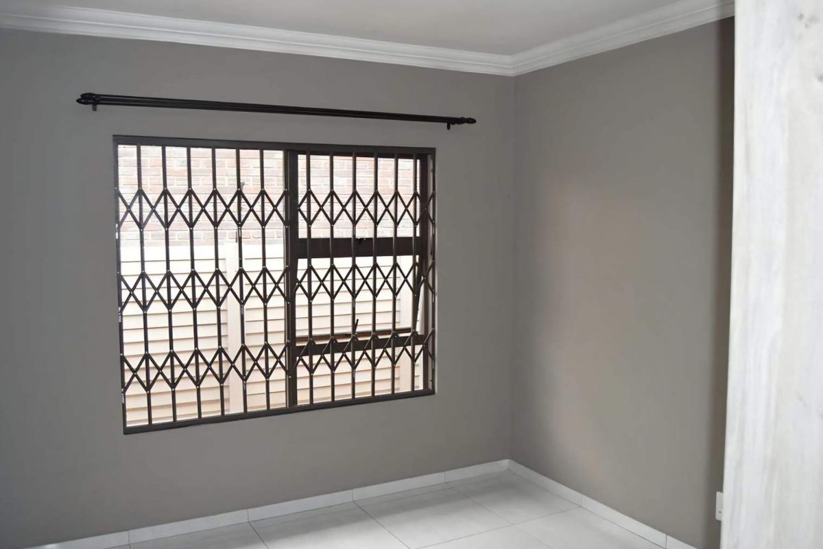 A clean, empty bedroom featuring neutral grey walls, white tiled flooring, and a window fitted with a security gate.