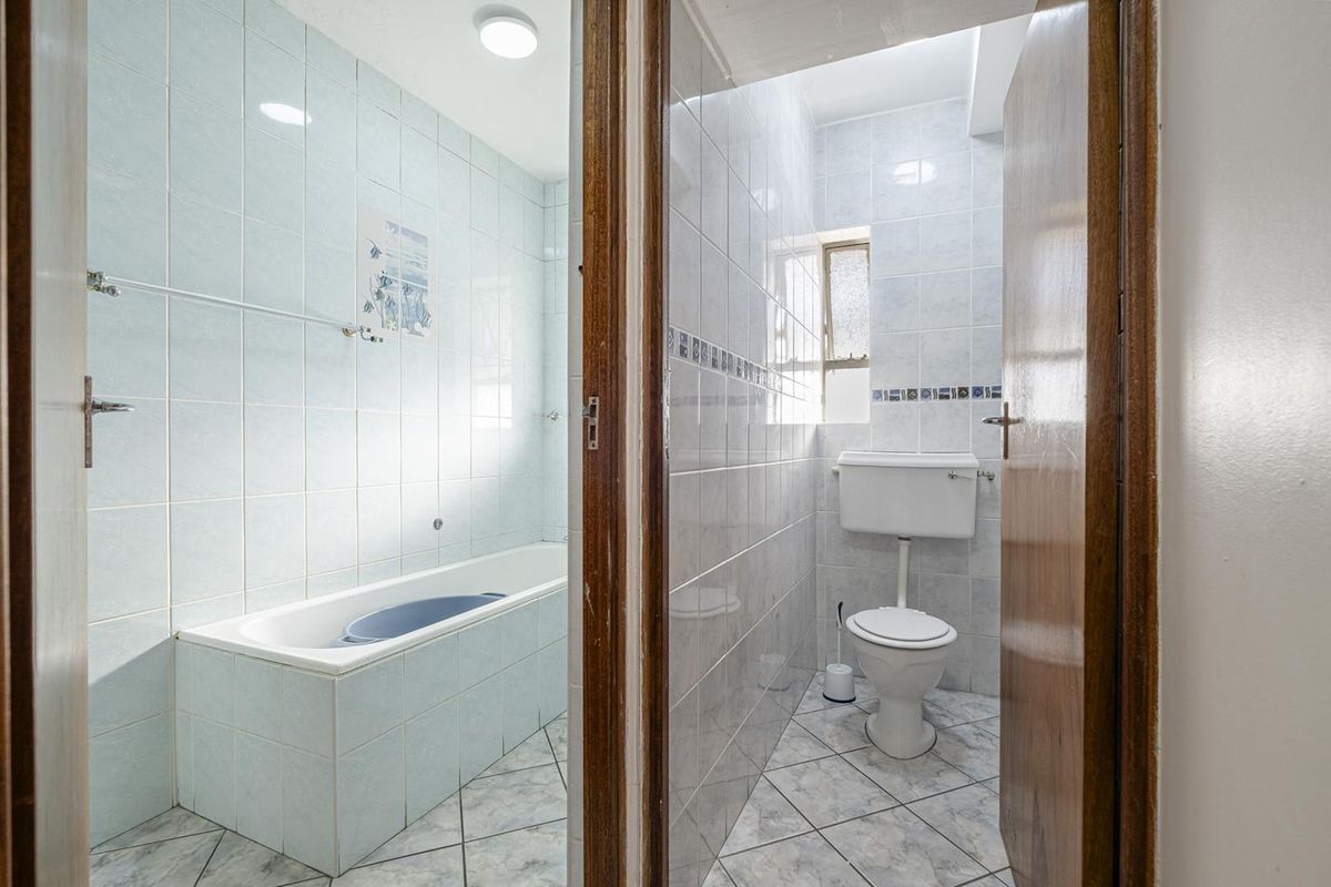 A functional bathroom featuring a built-in bathtub and a separate toilet area with light-colored ceramic tiling throughout.