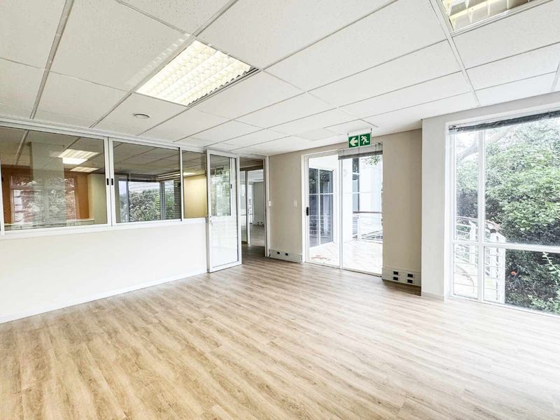 Bright and spacious open-plan office area featuring laminate flooring, suspended ceilings, glass partitions, and large windows providing natural light and garden views.