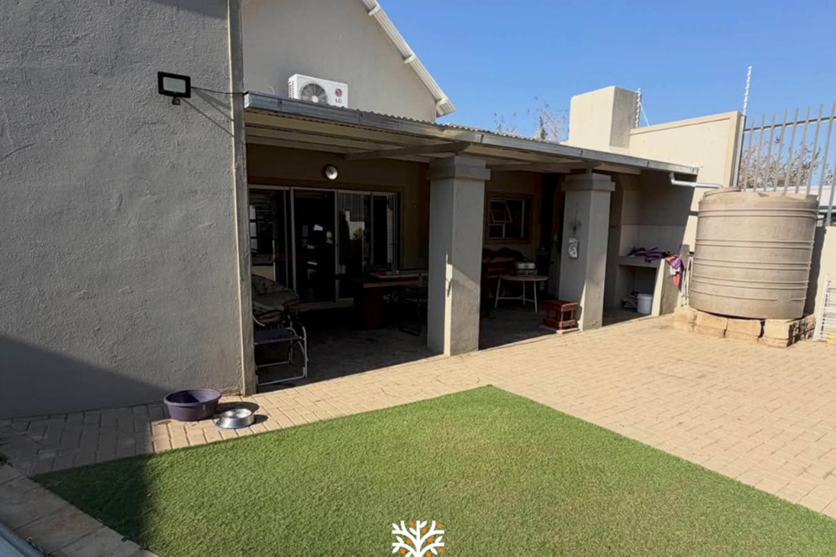 Rear exterior view featuring a covered patio, paved courtyard area, artificial lawn, and a water storage tank.