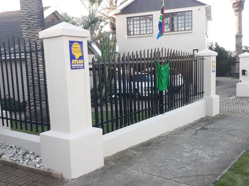 Front exterior view of the property featuring a secure perimeter wall with metal fencing, concrete pillars, and a paved driveway entrance.