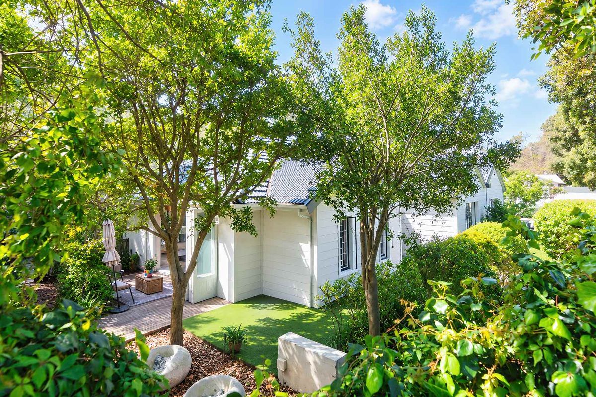 A charming rear exterior view featuring white weatherboard cladding, a corrugated metal roof, timber decking, and a well-maintained garden area.