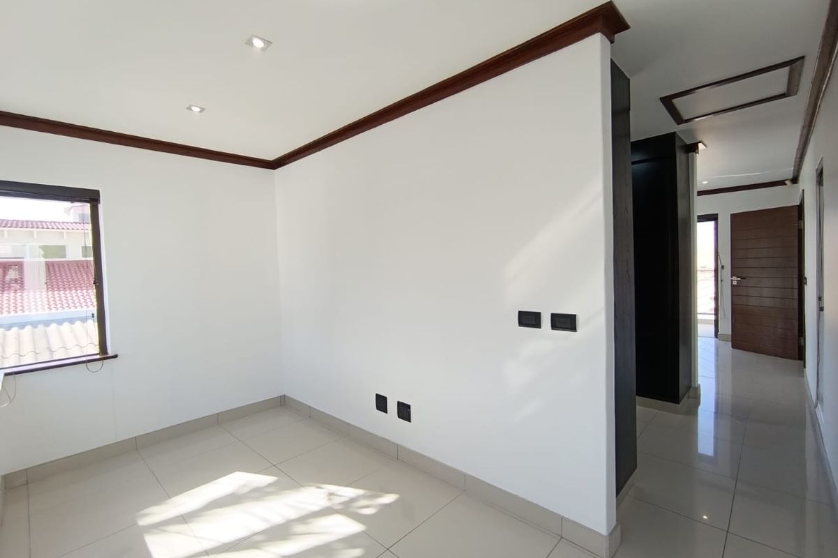A bright, empty bedroom featuring neutral tiled flooring, recessed ceiling lighting, and dark wood trim accents, leading into a hallway.
