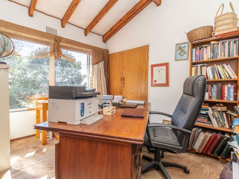 A home office featuring exposed timber ceiling beams, a built-in wooden cupboard, and a large window providing natural light.