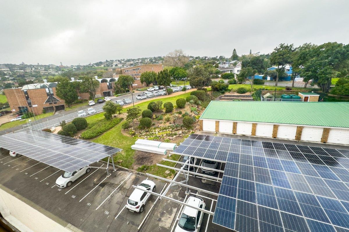 Elevated view overlooking a parking area equipped with solar panel carports, surrounding greenery, and residential buildings in the distance.