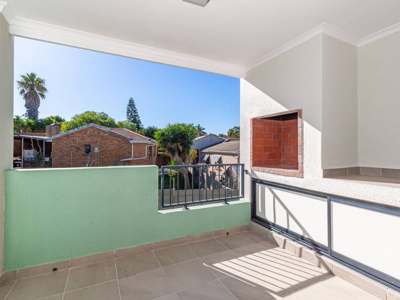 A clean, covered balcony featuring tiled flooring, a built-in brick braai, and metal balustrades, offering an open view of the surrounding neighborhood.