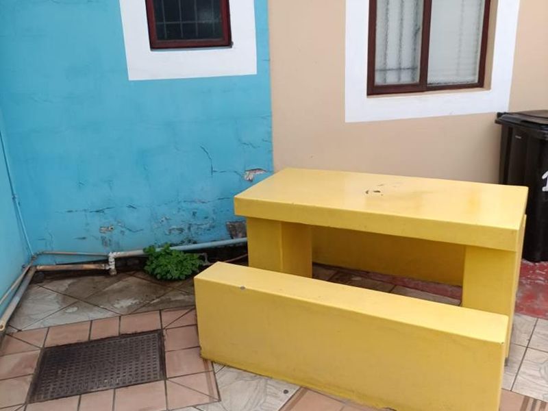 Outdoor patio area featuring built-in concrete seating and table, tiled flooring, and security-barred windows, requiring maintenance to the exterior wall finishes.