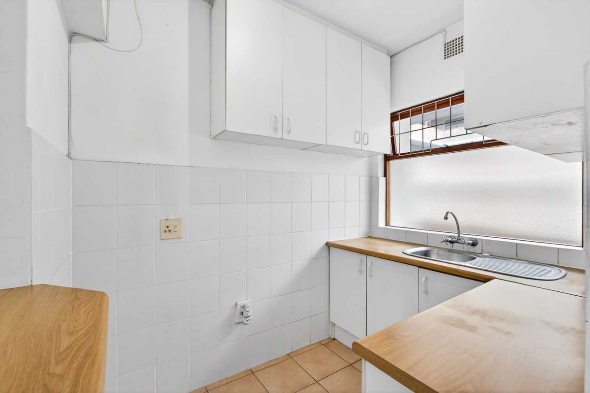 A functional, compact kitchen featuring white cabinetry, laminate countertops, a stainless steel sink, and tiled splashbacks, offering a clean canvas for potential updates.