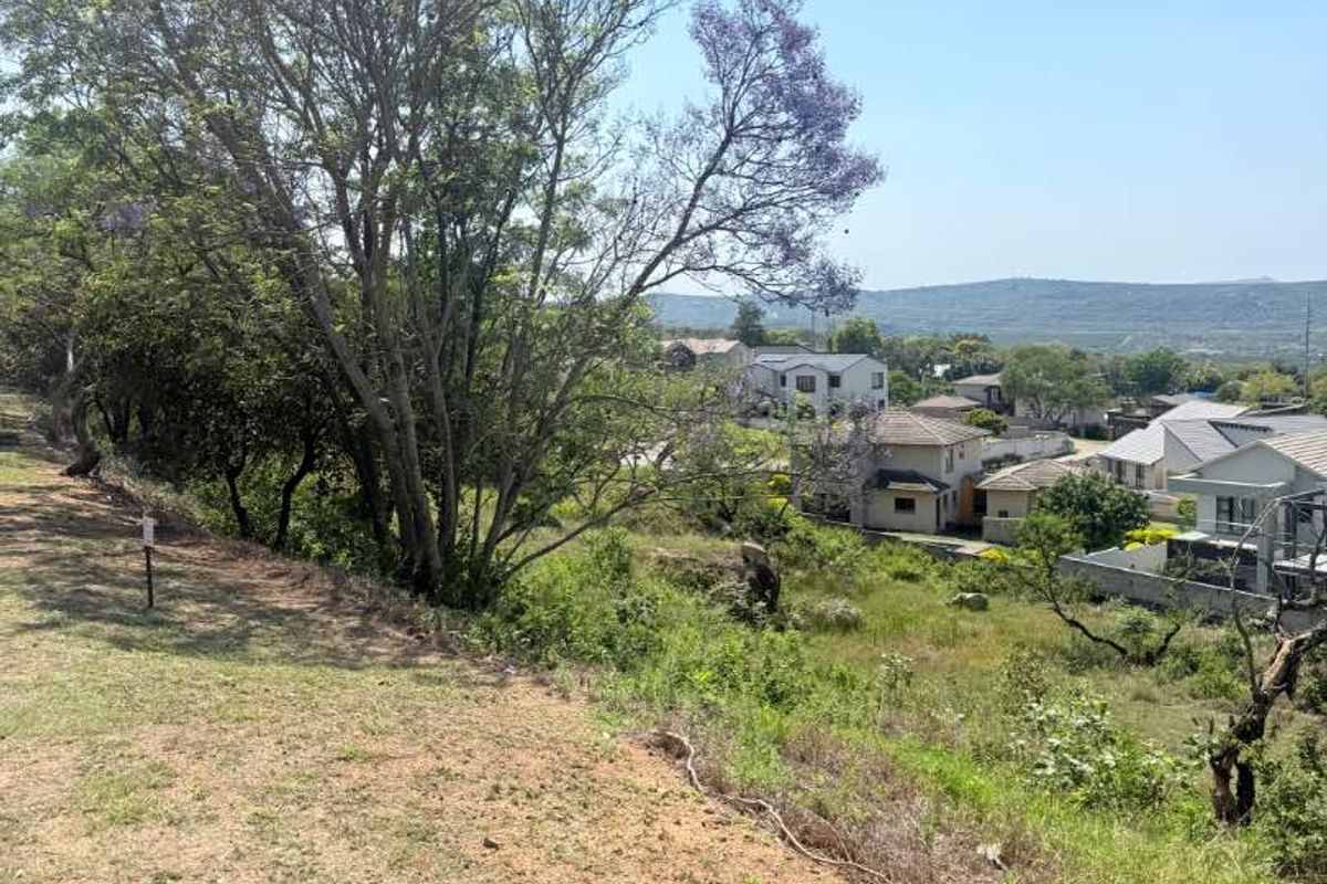 A view overlooking a sloped residential plot with mature trees and natural vegetation, showcasing the surrounding neighborhood and distant landscape.