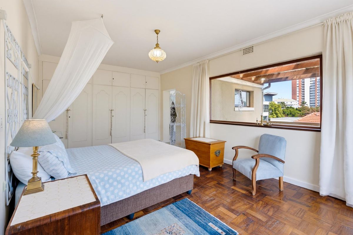 A well-maintained bedroom featuring classic parquet flooring, extensive built-in cupboard storage, and a large window providing natural light and exterior views.