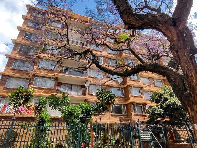 A multi-story apartment building featuring a brick facade, private balconies with metal railings, and secure perimeter fencing with an integrated security gate.