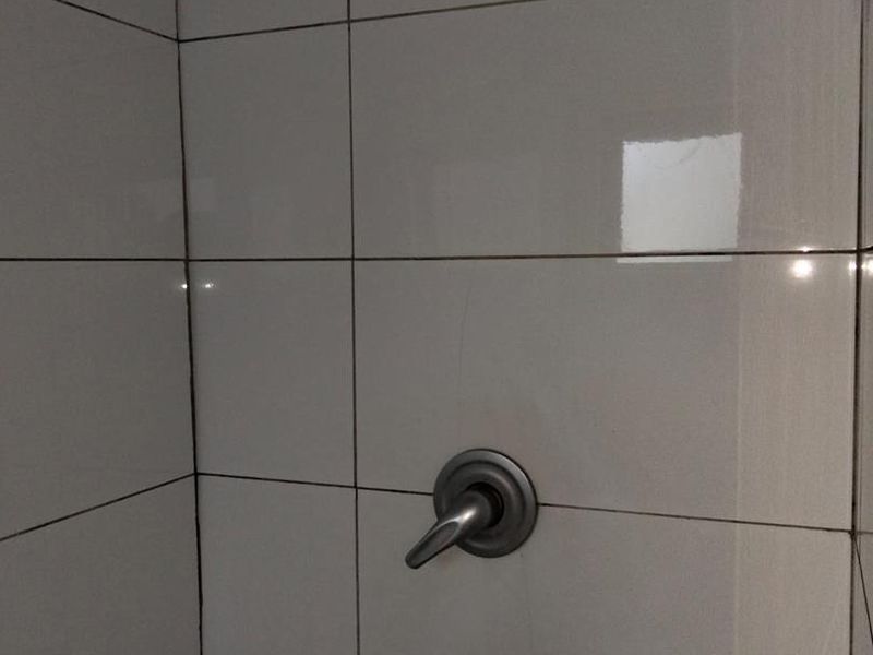 A close-up view of a shower enclosure featuring white ceramic wall tiles, a standard shower head, and a single-lever mixer tap.