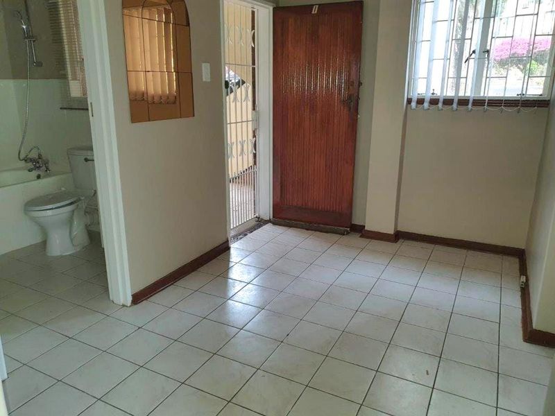 An empty living area featuring tiled flooring, a wooden entrance door, and security bars on the window, with an adjacent bathroom visible.