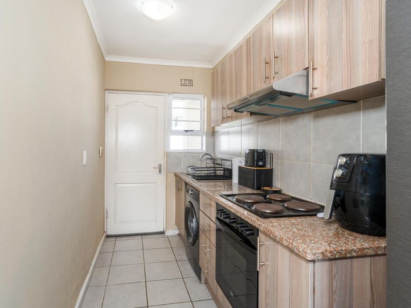 Compact kitchen featuring wood-finish cabinetry, granite countertops, built-in oven, electric hob, and tiled flooring with direct access to an exterior door.