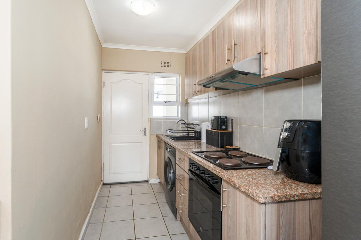 Compact kitchen featuring wood-finish cabinetry, granite countertops, built-in oven, electric hob, and tiled flooring with direct access to an exterior door.