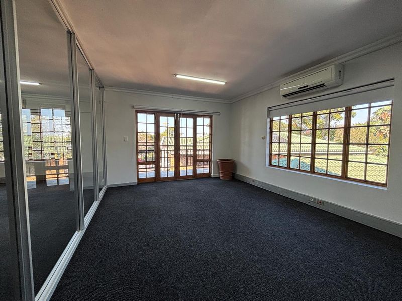 A spacious, empty office area featuring dark carpeting, a split-system air conditioner, large windows, and a mirrored sliding partition wall.