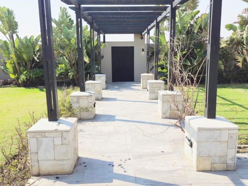 A paved outdoor walkway featuring a timber pergola structure supported by stone-clad pillars, leading to a private entrance in a landscaped garden setting.