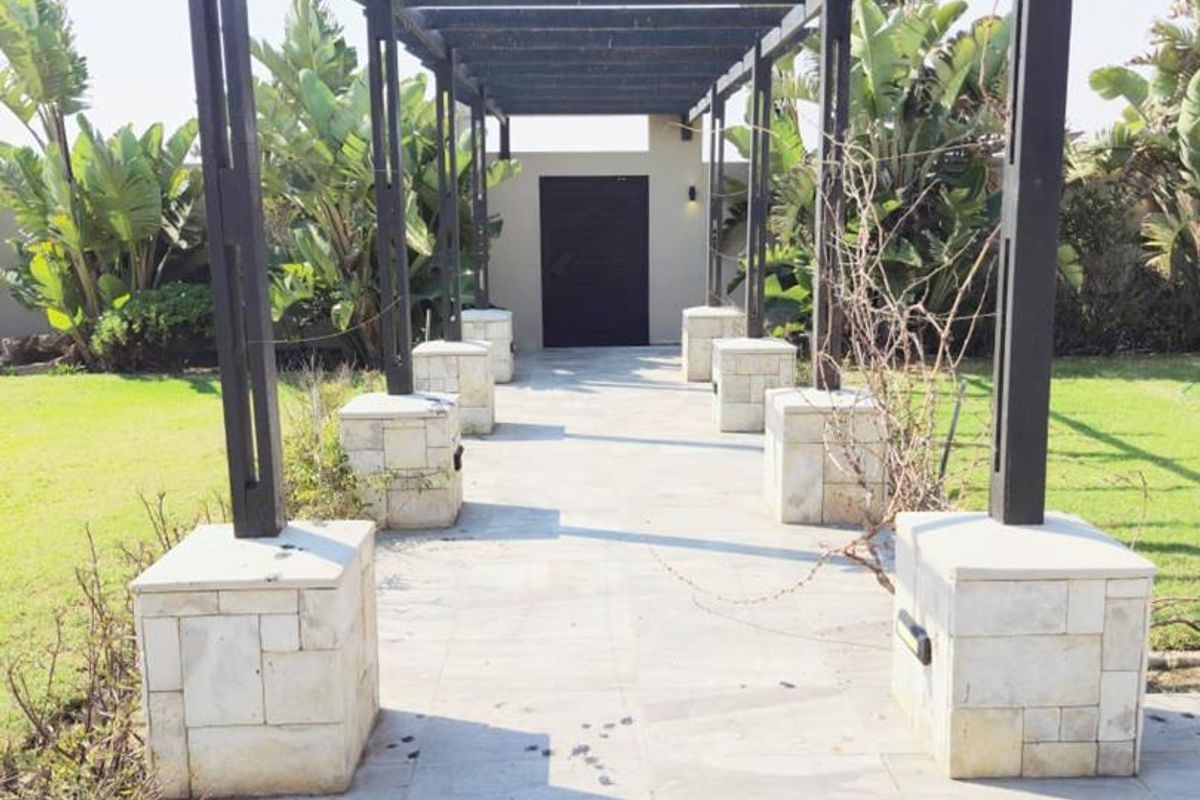 A paved outdoor walkway featuring a timber pergola structure supported by stone-clad pillars, leading to a private entrance in a landscaped garden setting.