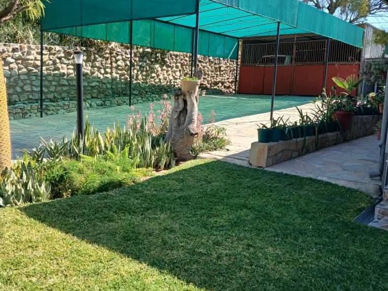 Rear garden area featuring a lawn, stone retaining wall, and a large green shade cloth carport structure over a paved driveway.