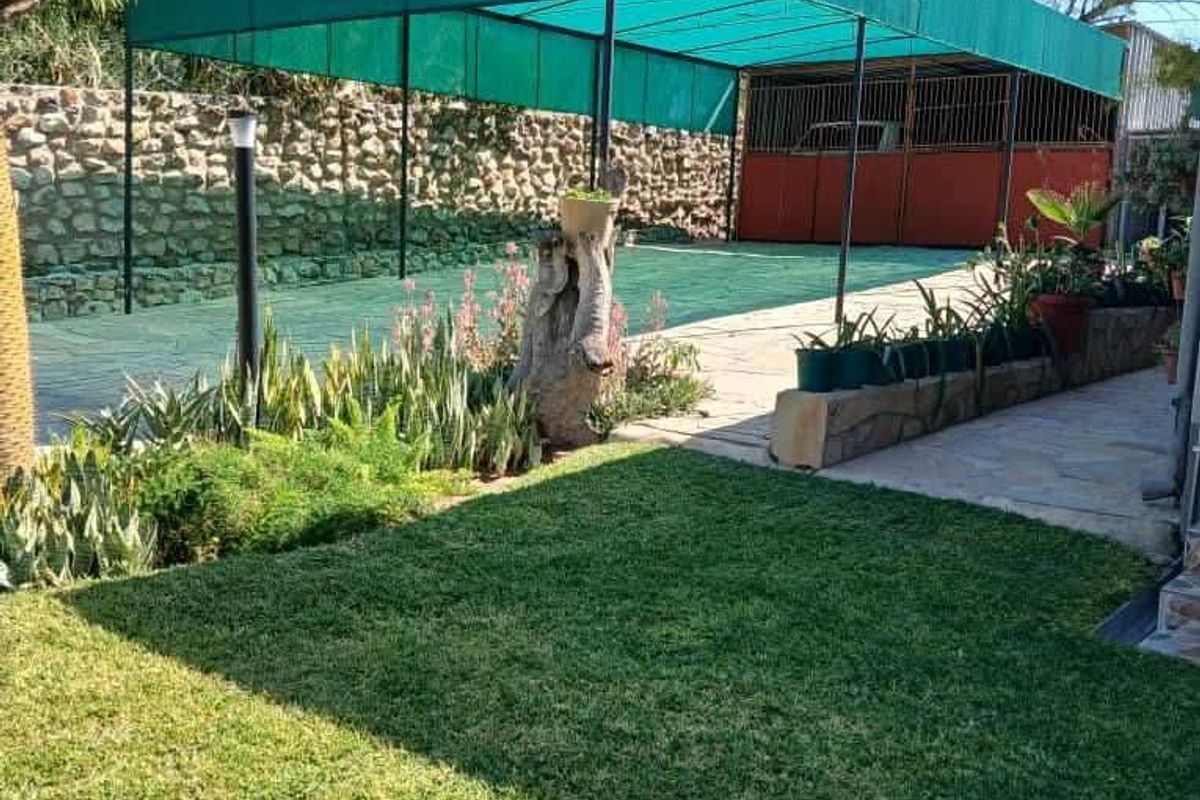 Rear garden area featuring a lawn, stone retaining wall, and a large green shade cloth carport structure over a paved driveway.