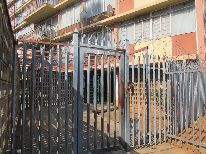 Exterior view of an apartment building entrance featuring a secure metal gate, perimeter fencing with razor wire, and multi-story brick facade.