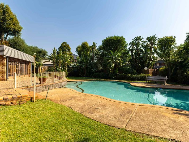 A spacious outdoor pool area featuring a curved swimming pool, paved surround, and an adjacent brick patio, set within a mature landscaped garden.