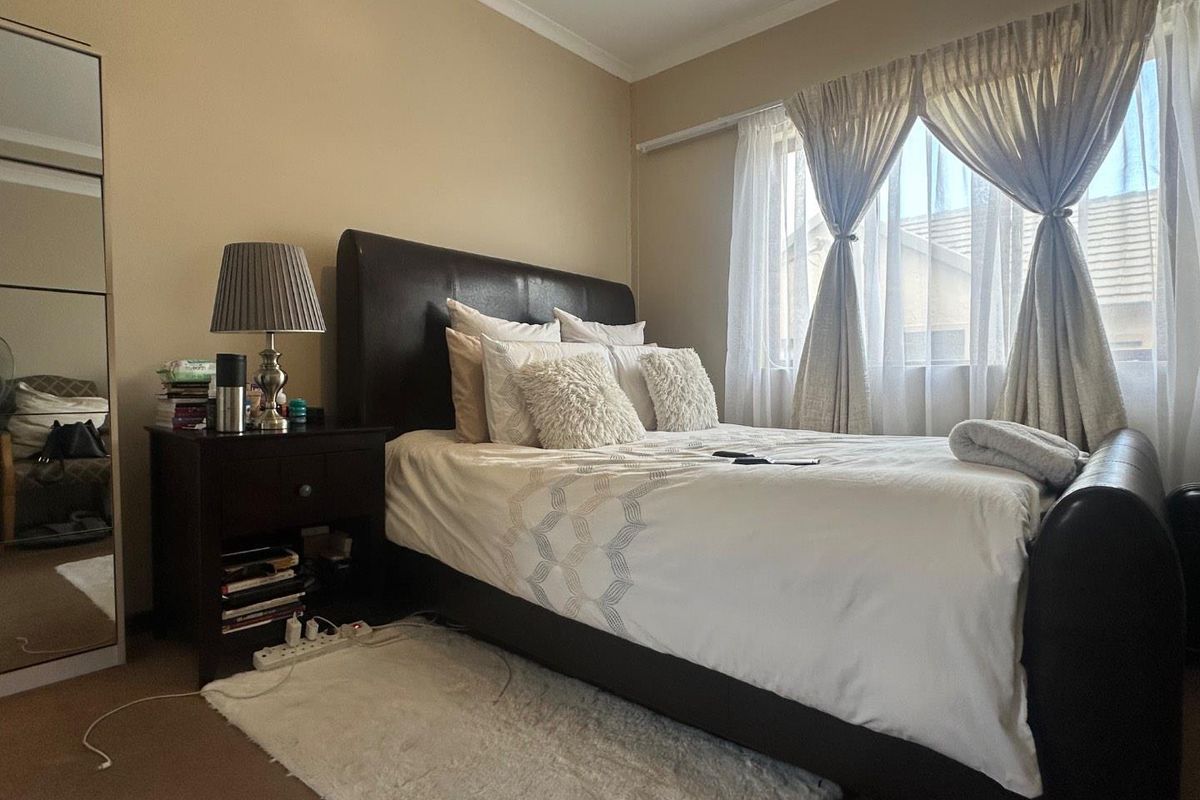 A well-lit bedroom featuring neutral wall tones, carpeted flooring, and a window with fitted curtain tracks.