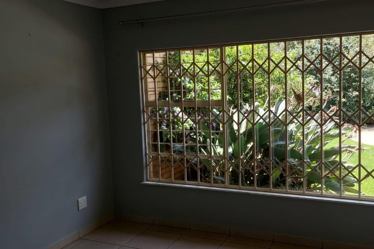A bright, empty bedroom featuring tiled flooring and a large window fitted with security bars, offering a view of the garden and pool area.