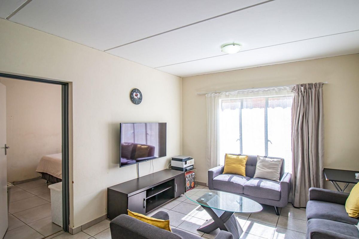 A functional living room featuring tiled flooring, large windows for natural light, and direct access to an adjacent bedroom.
