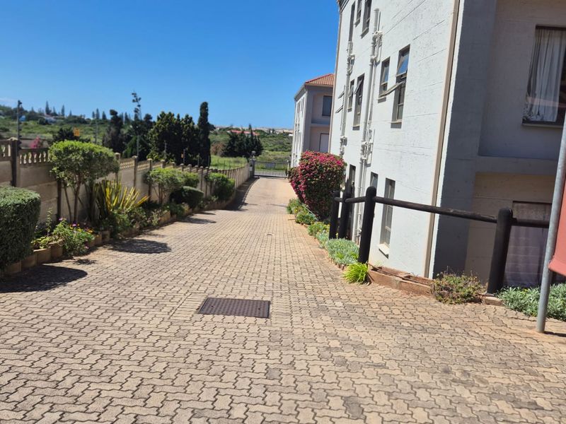 A paved driveway leading to the apartment complex entrance, featuring boundary walls, landscaped garden borders, and clear access to the building facade.