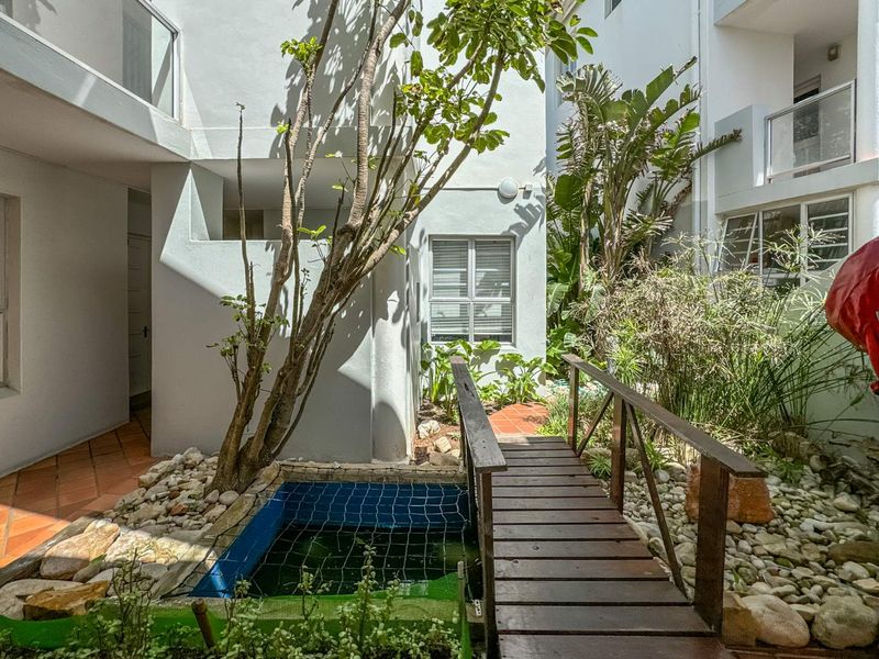 A quiet courtyard area featuring a small pond, a wooden footbridge, and terracotta paving surrounded by established garden greenery.