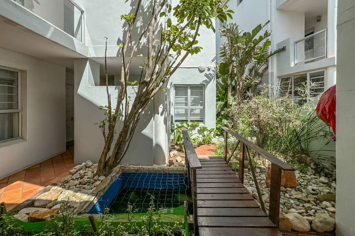 A quiet courtyard area featuring a small pond, a wooden footbridge, and terracotta paving surrounded by established garden greenery.