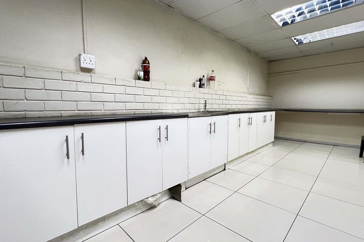 A functional industrial kitchenette featuring white cabinetry, a dark countertop, a stainless steel sink, and tiled flooring with a suspended ceiling.