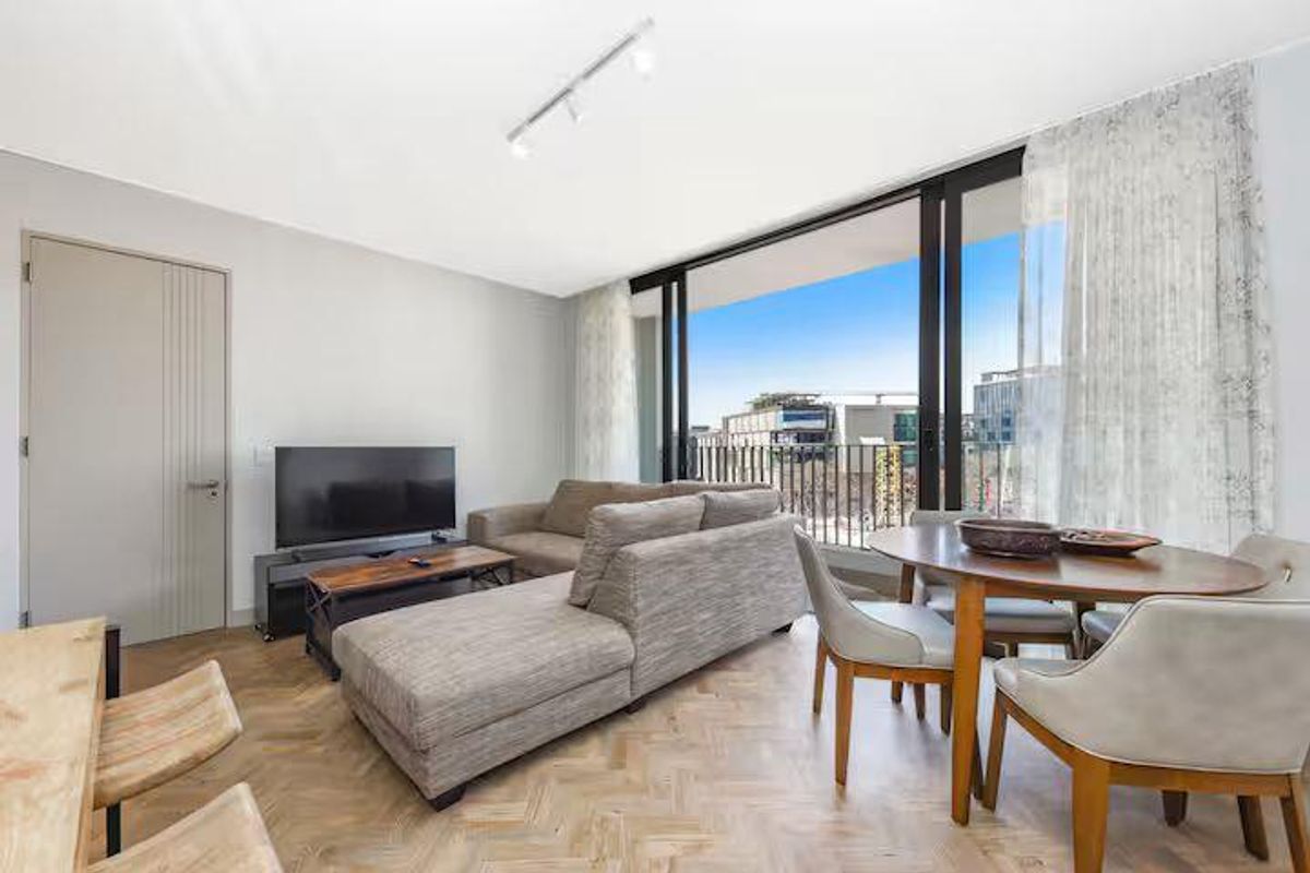 Open-plan living and dining area featuring herringbone timber flooring, modern track lighting, and large sliding glass doors opening to a balcony with urban views.