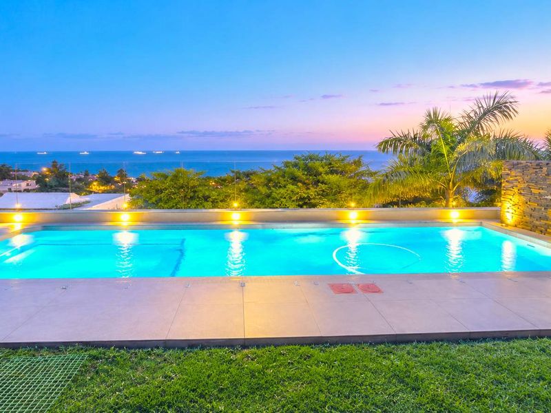 A beautifully illuminated swimming pool area featuring a stone accent wall, glass balustrade, and stunning ocean views at sunset.