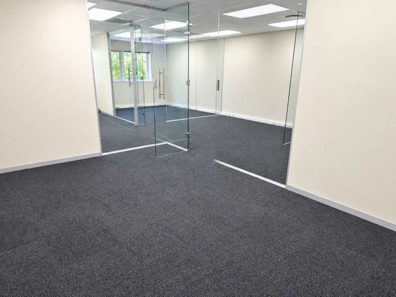 A modern, vacant office space featuring glass partition walls, a suspended ceiling with recessed lighting, and clean carpet flooring.