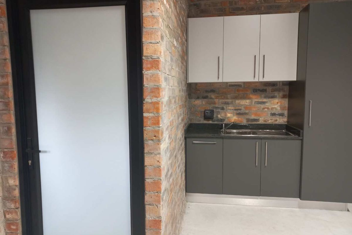 A compact kitchenette featuring modern grey cabinetry, a stainless steel sink, and a striking exposed brick wall finish.