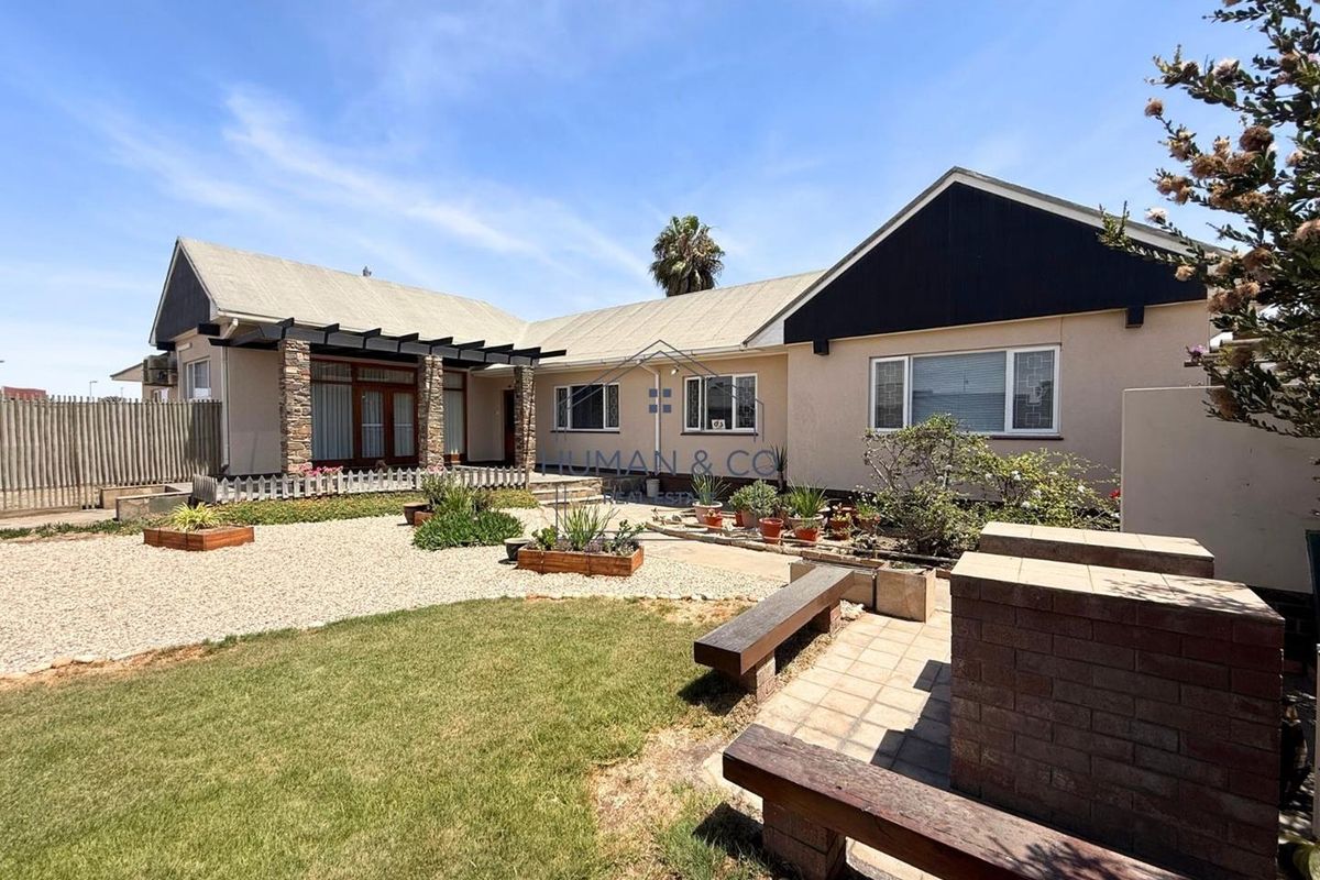 A spacious rear exterior view featuring a landscaped garden, paved patio area with built-in brick seating, and a covered pergola structure.